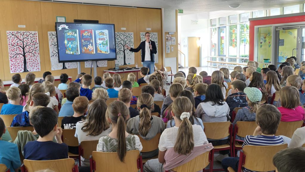 Ein Mann steht vor einer Gruppe von Kinder und präsentiert auf einen Bildschirm Kinderbücher